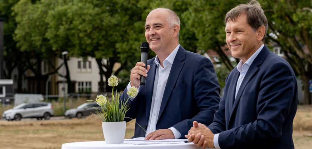 Der Geschäftsführer der SEG Herne und der Oberbürgermeister von Herne stehen lachend an einem Stehtisch vor Bäumen und halten eine Rede