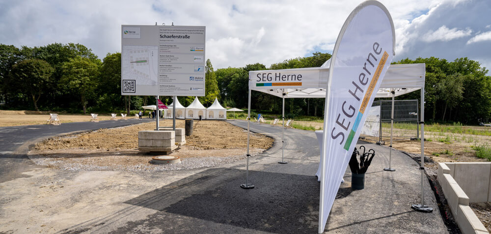 Eine Beachflag mit dem Schriftzug "SEG Herne" und ein großes Baustellenschild mit Infos zum Projekt markieren den Eingang zum Grundstück für das Bauvorhaben an der Schäferstraße in Herne